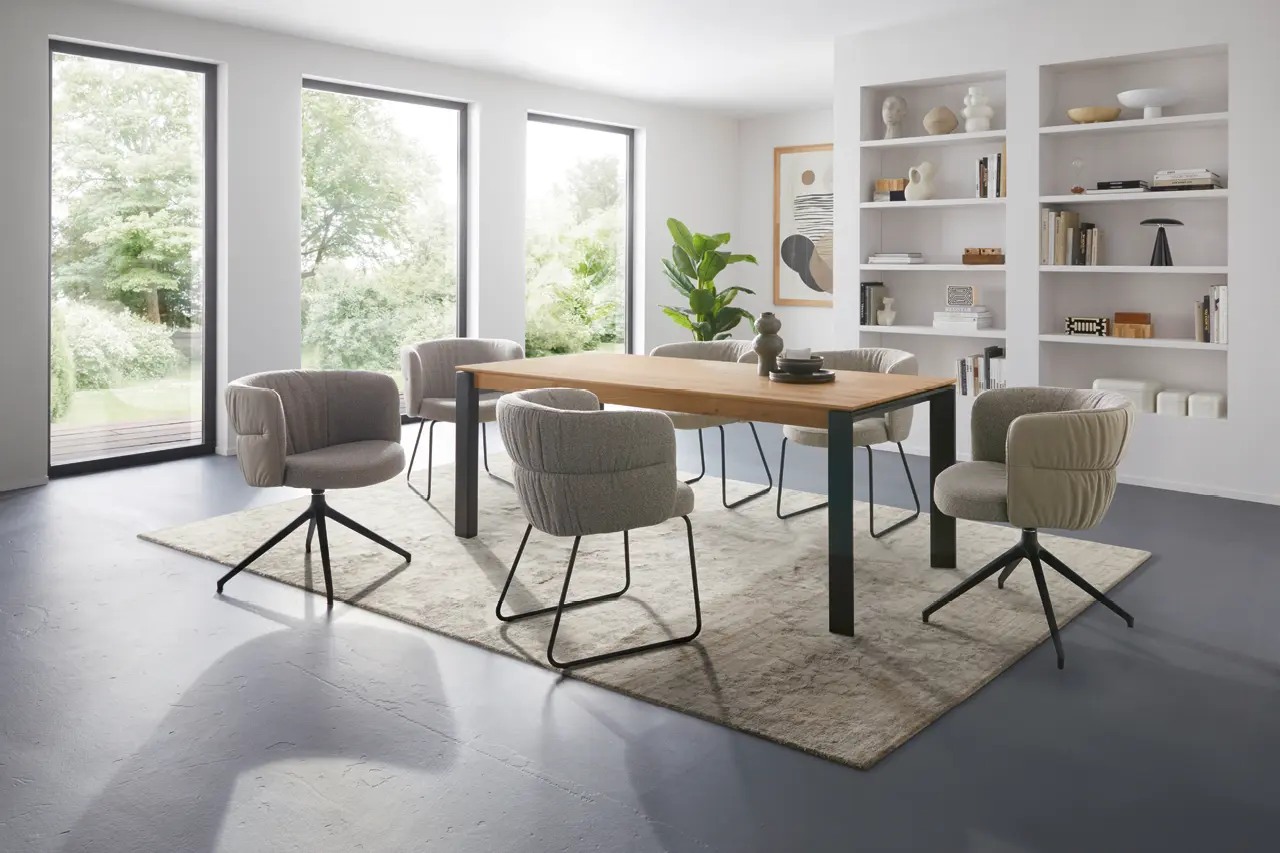 Helles Esszimmer mit großem Esstisch aus Holz und modernen Polsterstühlen in Grau. Stilvolle Einrichtung mit Teppich, Regalwand und großen Fenstern, ideal für modernes Wohnen und gemütliche Familienessen im zeitlosen Design.